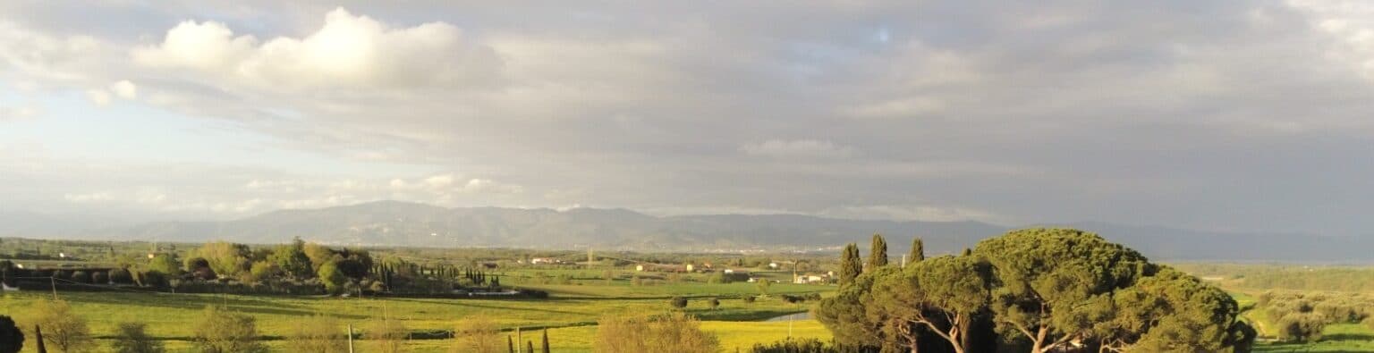 Vista aérea de la llanura cultivada entre las colinas, con hileras de cipreses y pinos, nubes bajas y las montañas de los Apeninos difuminadas en el horizonte