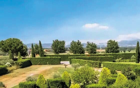 Eine sauber geschnittene Buchsbaumhecke im Vordergrund, Zypressen und vereinzelte Bäume in mittlerer Höhe, offene Felder und verschwommene Hügel am Horizont unter blauem Himmel mit einer weißen Wolke