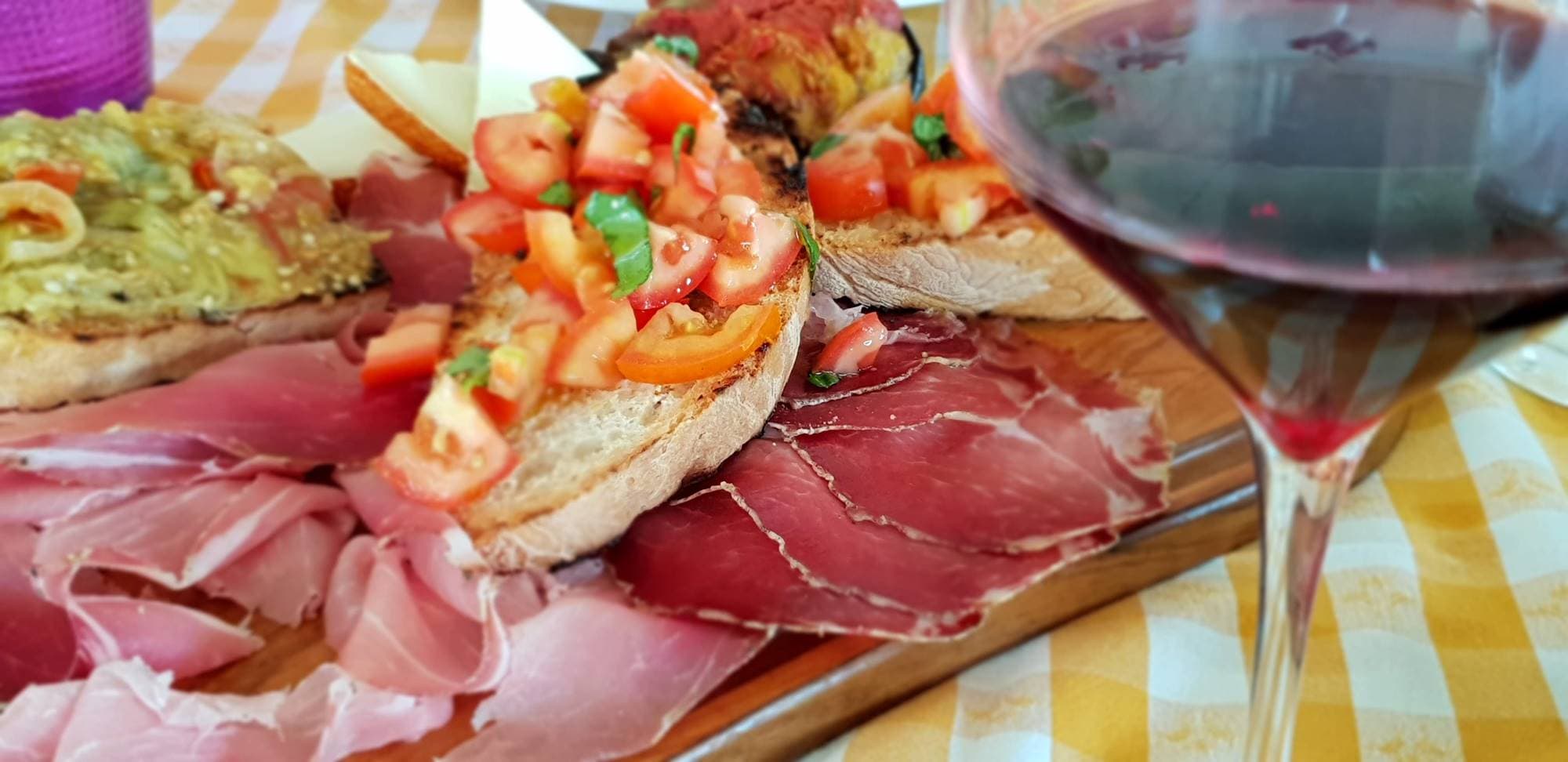 Tagliere di prosciutto e bruschette al pomodoro accanto a un bicchiere di vino rosso, su una tovaglia a quadri