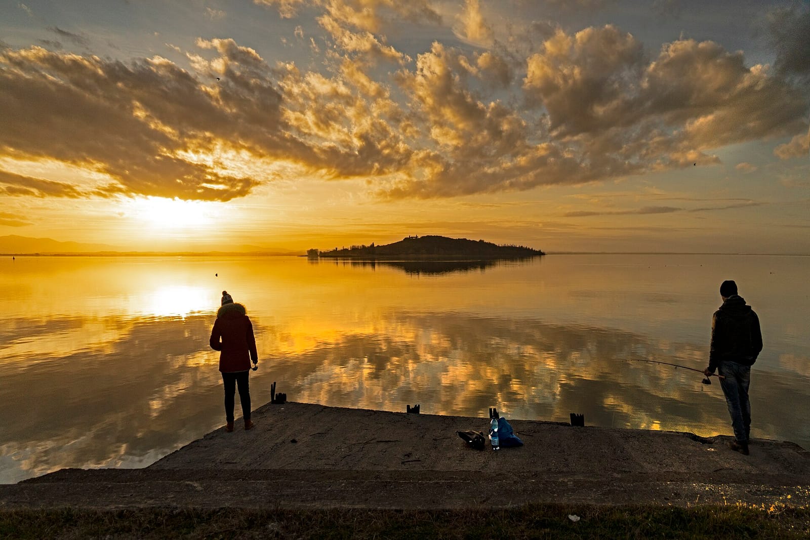 Isola Polvese at sunset seen from the shore, two anglers silhouetted on a slipway